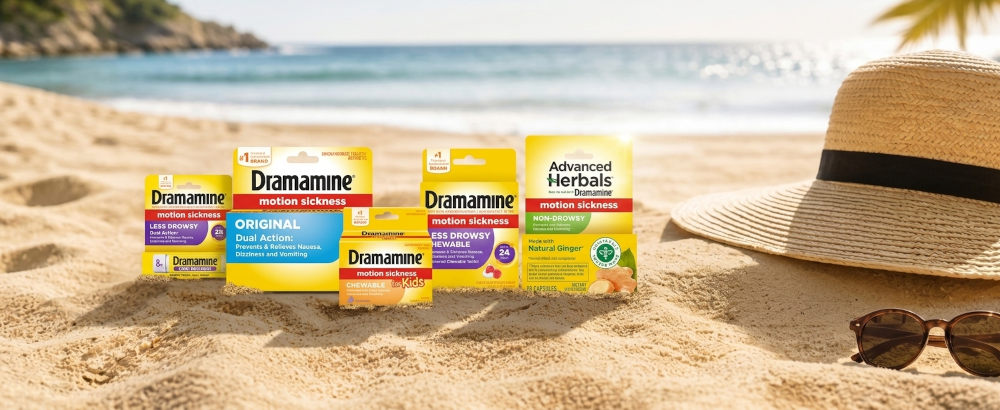 Dramamine motion sickness tablet packages sit on sunny beach sand beside a straw hat and sunglasses, with ocean waves behind them, evoking carefree travel.