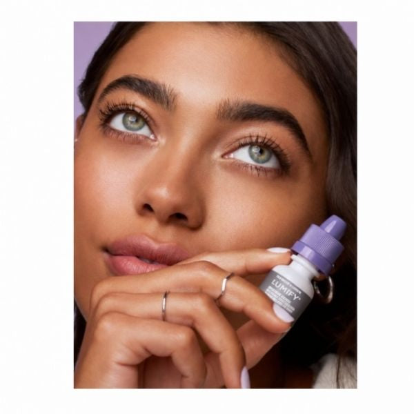 Close-up of a woman holding a small white and purple Lumify eye drops dropper bottle near her face, with a purple background.